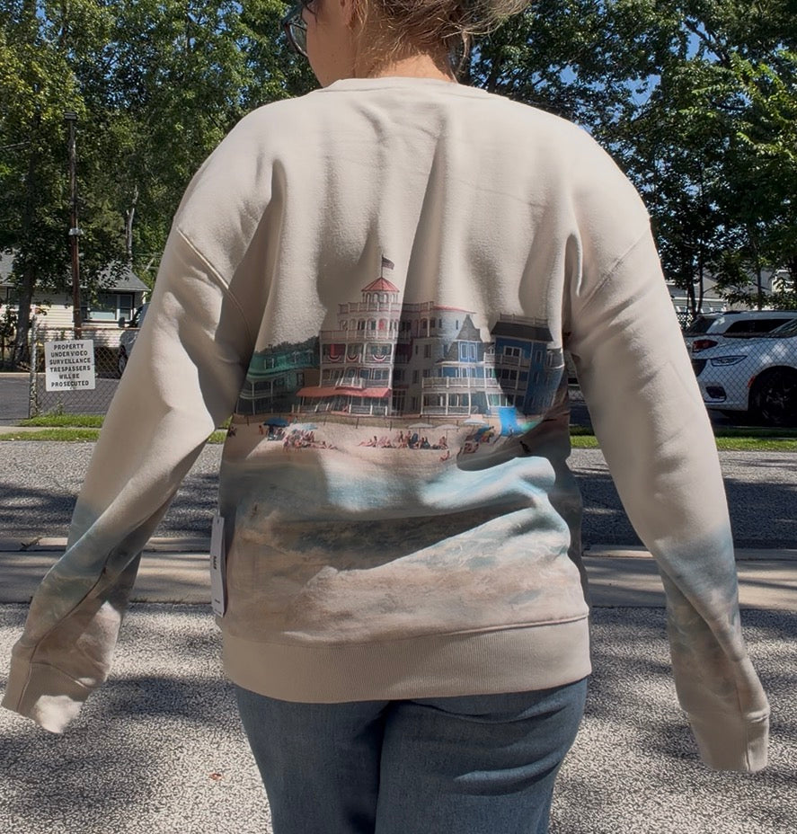 A person viewed from behind wearing a beige sweatshirt with a large printed beach house and shoreline scene across the back, paired with blue jeans outdoors on a paved area with trees and a car in the background.