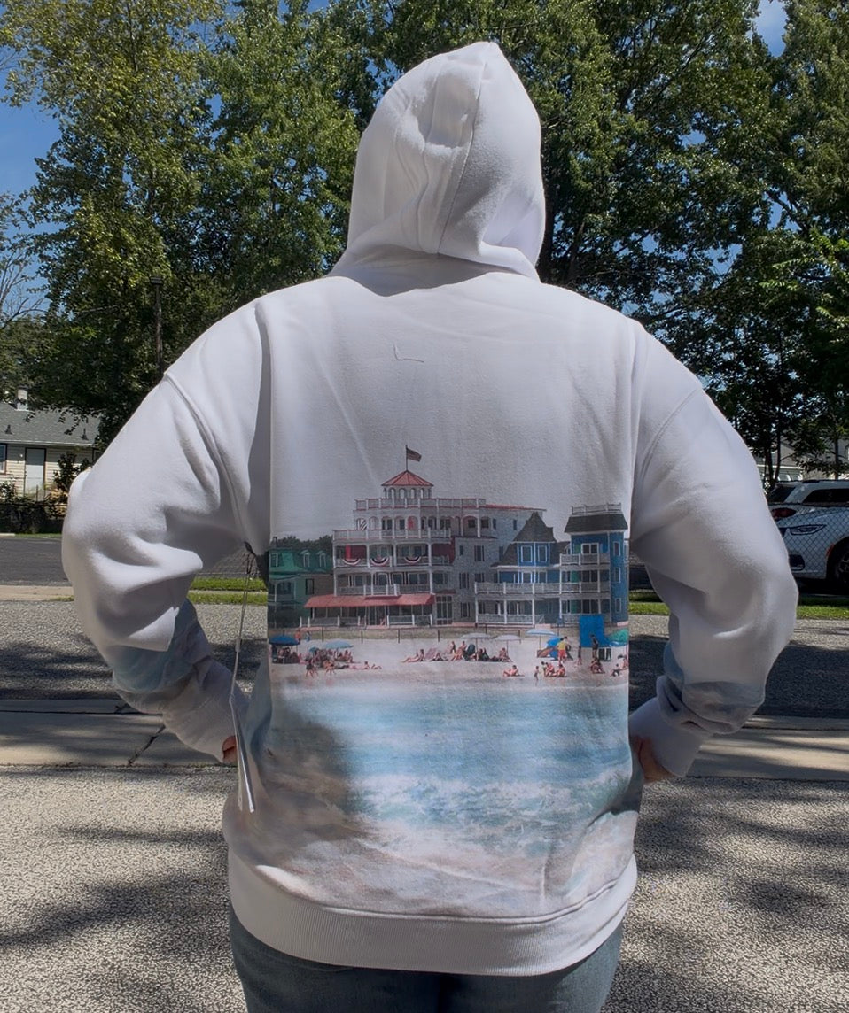 A person standing outdoors with their back to the camera, wearing a white hoodie featuring a large printed beach resort scene on the back.