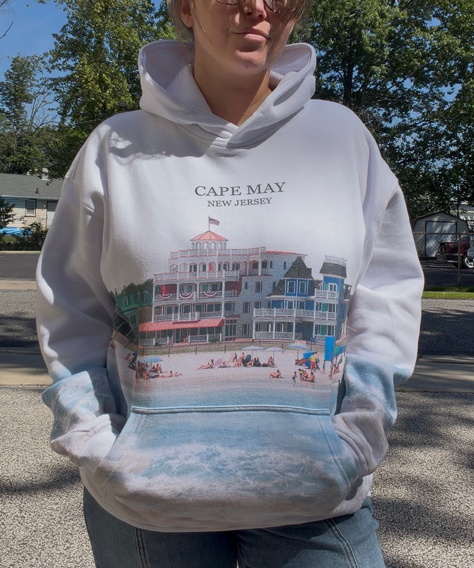 A person wearing a white pullover hoodie with a large Cape May, New Jersey beachfront building graphic and text on the front, photographed outdoors on a sunny day.