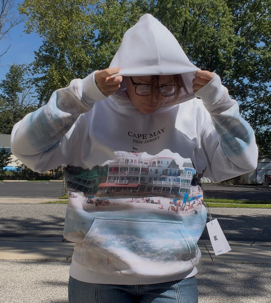 A person wearing a white Cape May, New Jersey themed hoodie with a beach scene graphic on the front, standing outdoors on a sunny day.