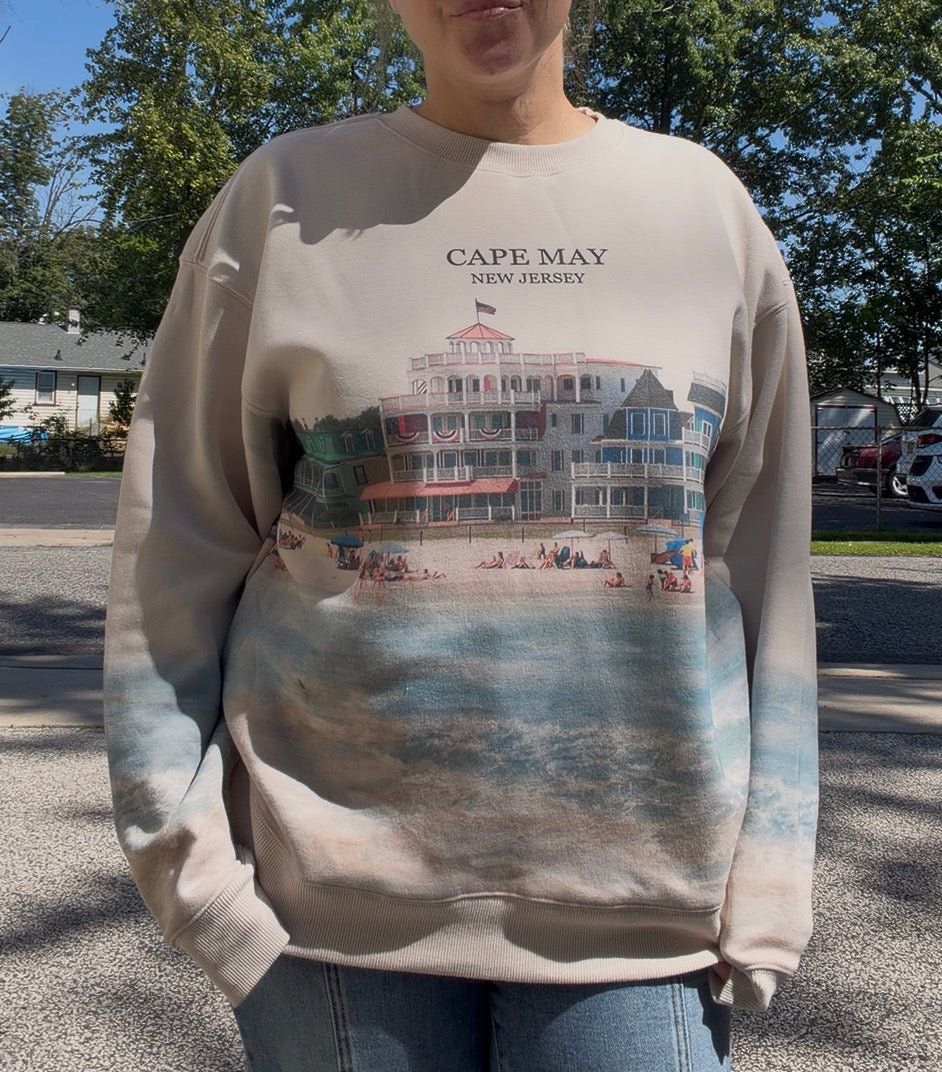 A person wearing a beige crewneck sweatshirt with a graphic print of a Cape May New Jersey beachfront building and water scene. The sweatshirt features a gradient transition from beige at the top to blue and gray at the bottom.