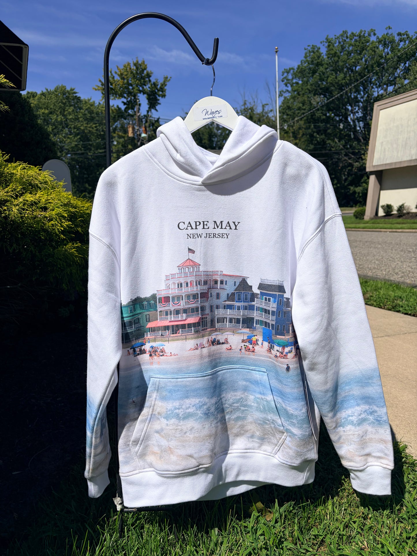 A white pullover hoodie hanging on a black metal hanger outdoors on a grassy lawn. The hoodie features a graphic print of a beach boardwalk building and ocean waves, with the text 'CAPE MAY NEW JERSEY' printed above the illustration.