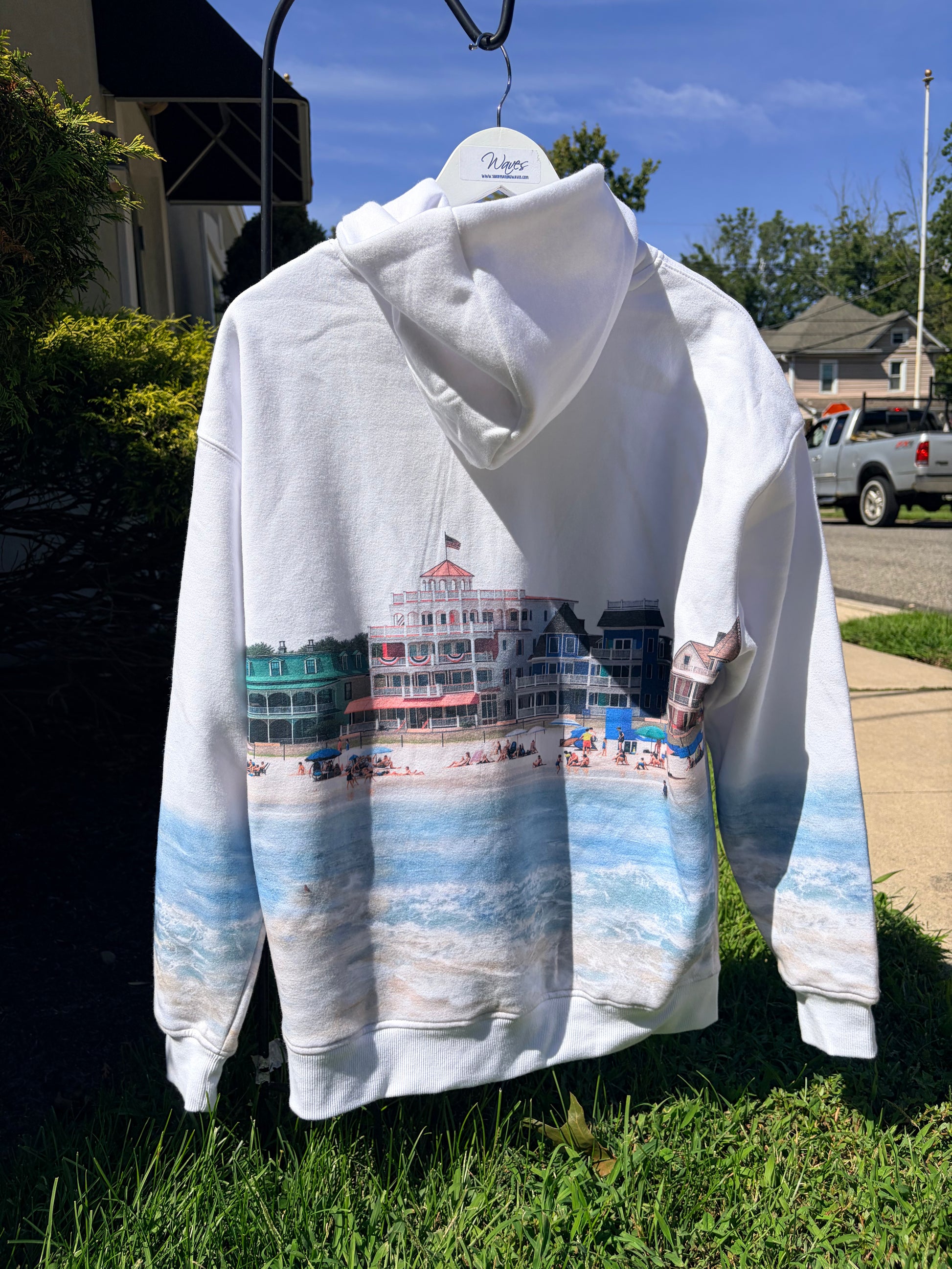 A white pullover hoodie hanging outdoors on a hanger, featuring a printed beach and boardwalk scene with a large red-and-white building and blue ocean waves. The hoodie is photographed in a real outdoor setting with grass, trees, houses, and a parked truck visible.