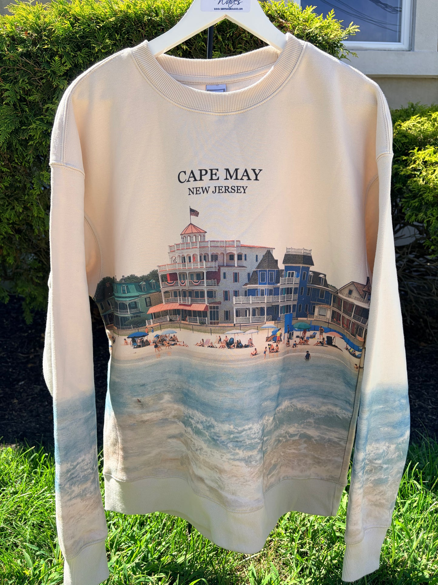 A cream-colored long-sleeve sweatshirt hanging on a white plastic hanger outdoors. The sweatshirt features a graphic print of a Cape May, New Jersey beach scene with beach houses and ocean waves, and the text 'CAPE MAY NEW JERSEY' printed above the graphic.