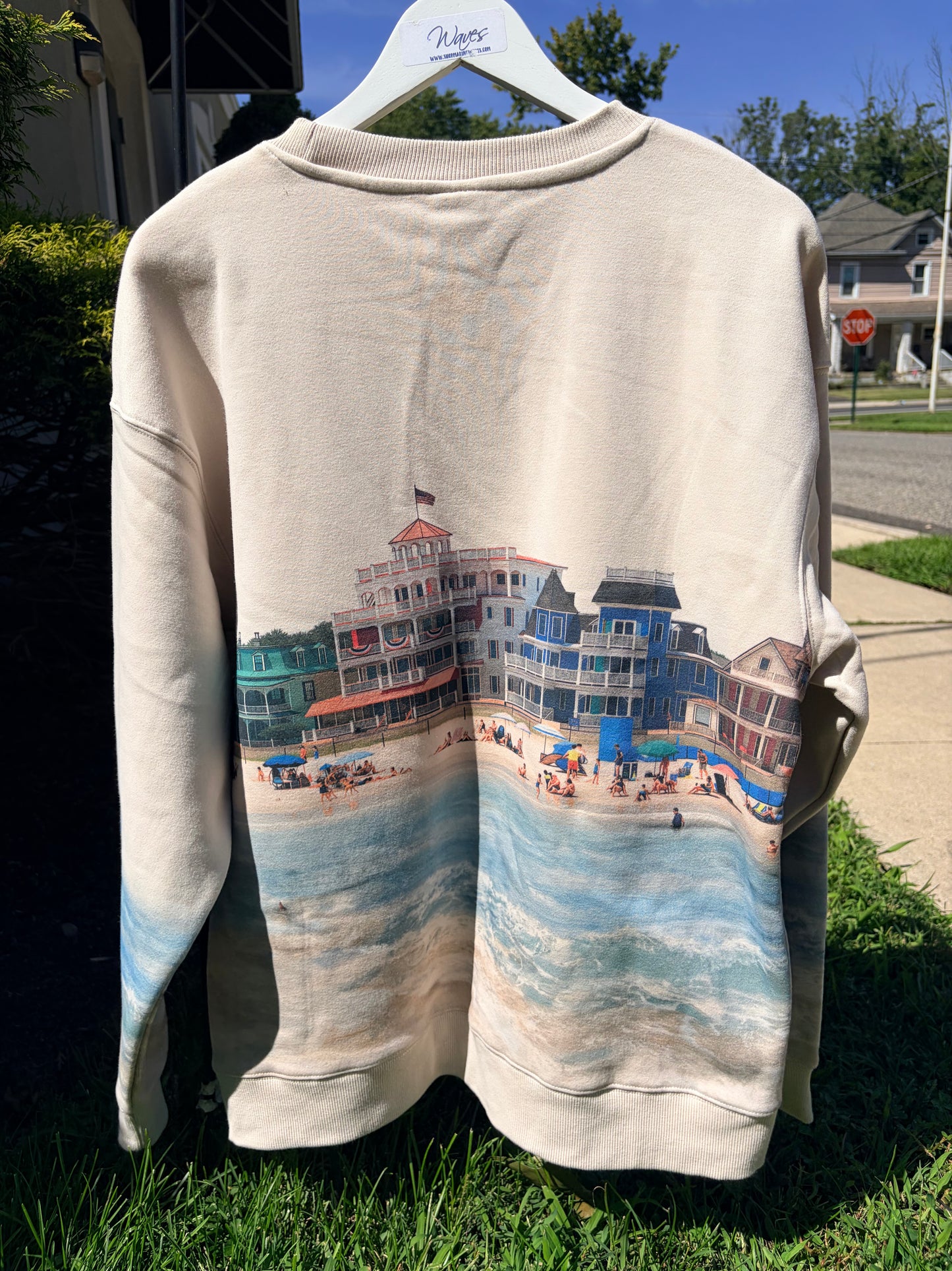 A cream-colored crewneck sweatshirt hanging on a white hanger outdoors on grass, featuring a large graphic print of colorful beach boardwalk houses and ocean waves across the front.