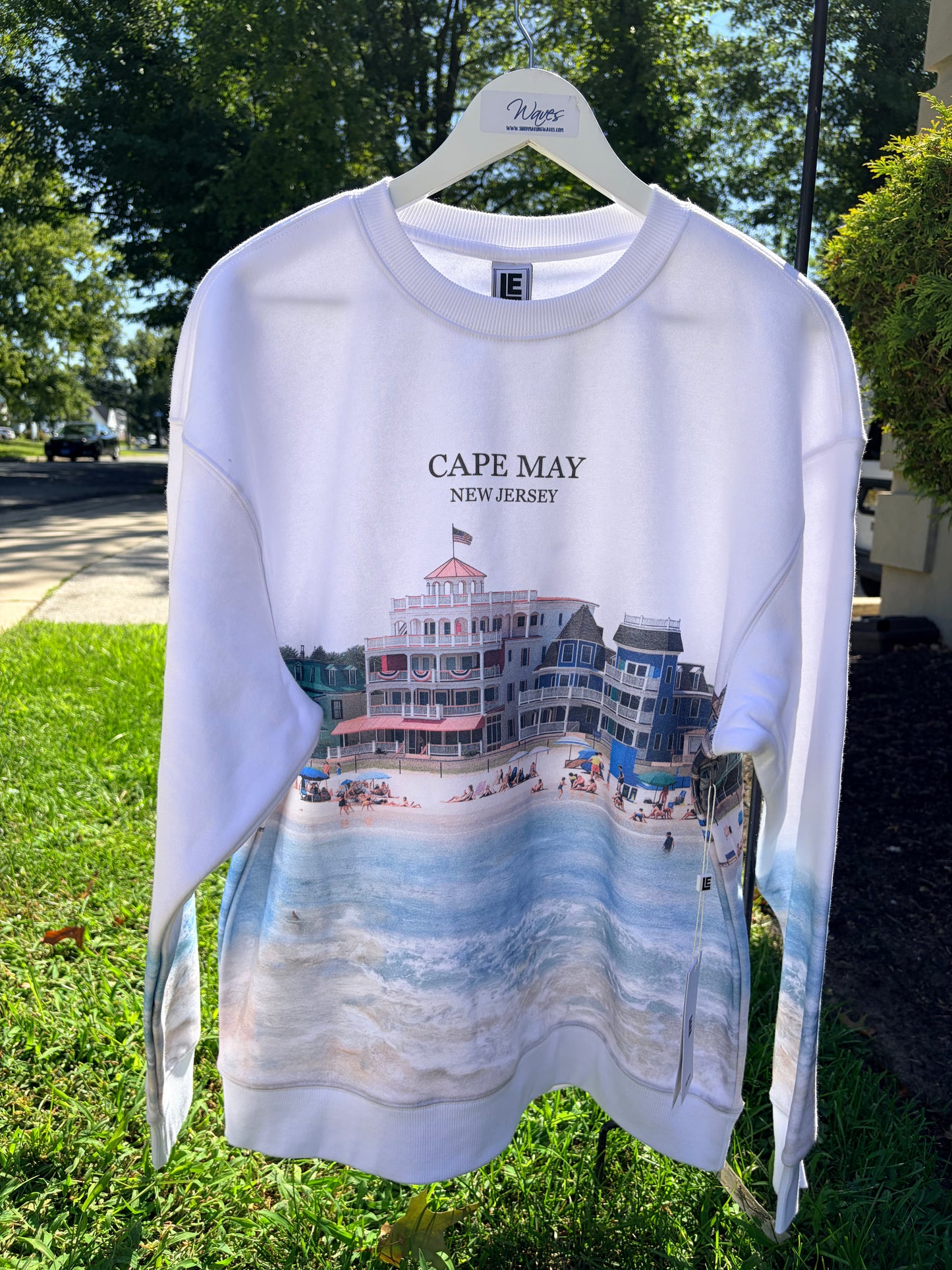 A white long-sleeve crewneck sweatshirt hanging on a white hanger outdoors on a grassy lawn. The sweatshirt features a graphic print of a beach boardwalk building with the text 'CAPE MAY NEW JERSEY' above a scenic beach and ocean print.