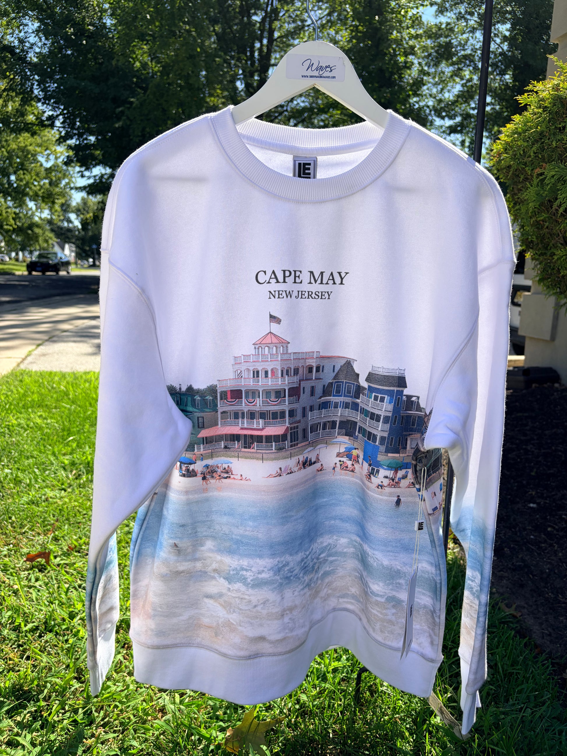 A white long-sleeve crewneck sweatshirt hanging on a white hanger outdoors on a grassy lawn. The sweatshirt features a graphic print of a beach boardwalk building with the text 'CAPE MAY NEW JERSEY' above a scenic beach and ocean print.