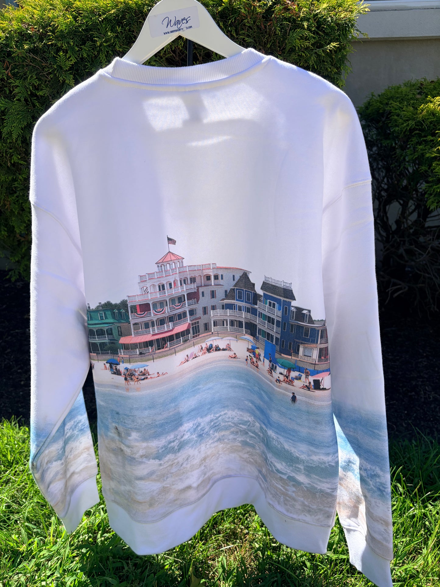 A white long-sleeve crewneck sweatshirt hanging on a white hanger outdoors. The back of the sweatshirt features a large graphic print of a beach boardwalk scene with colorful houses, a sandy beach, and blue ocean waves.