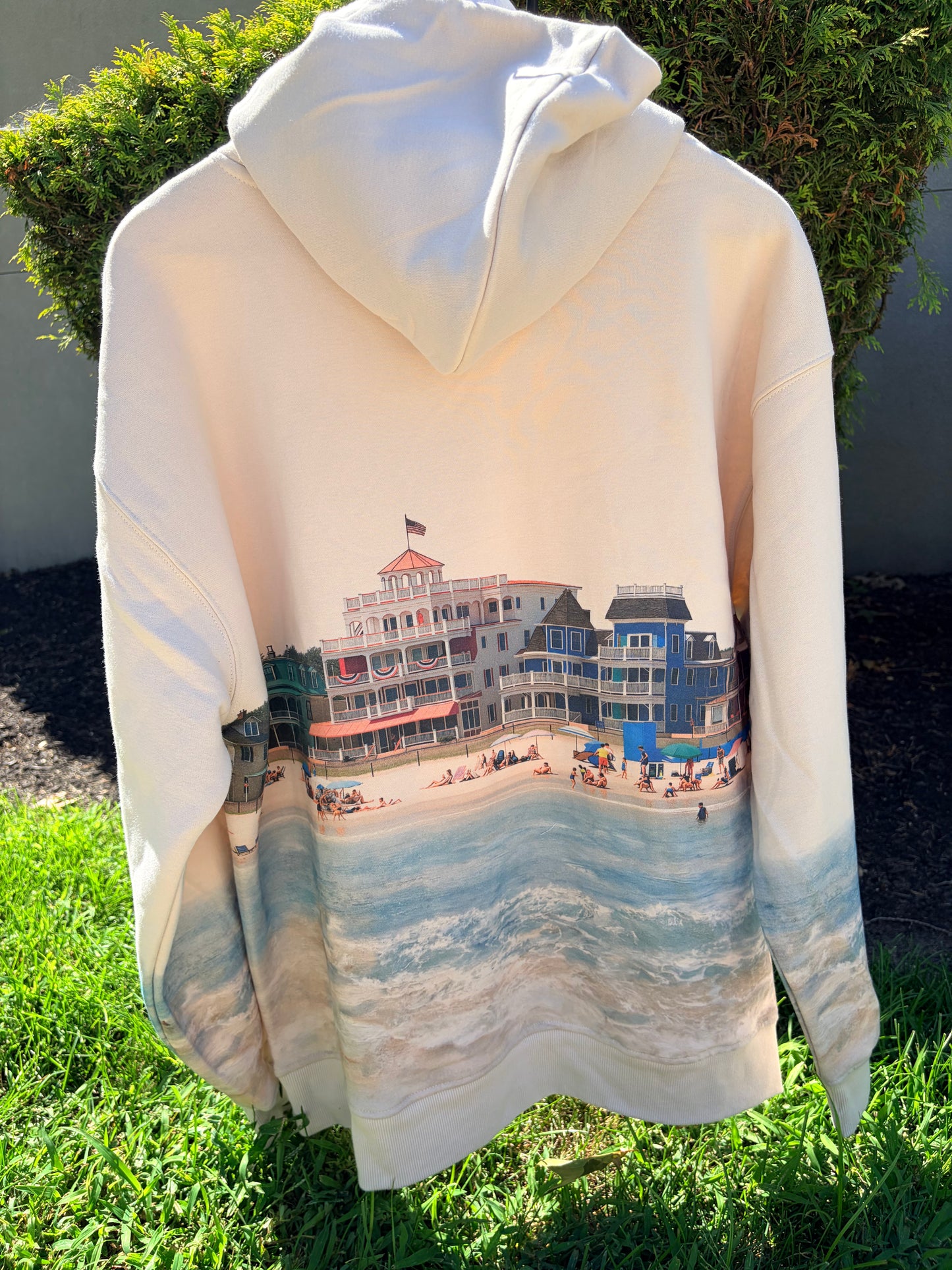 A cream-colored hooded sweatshirt with a large beach and boardwalk print on the back, displayed outdoors on green grass with shrubs in the background.