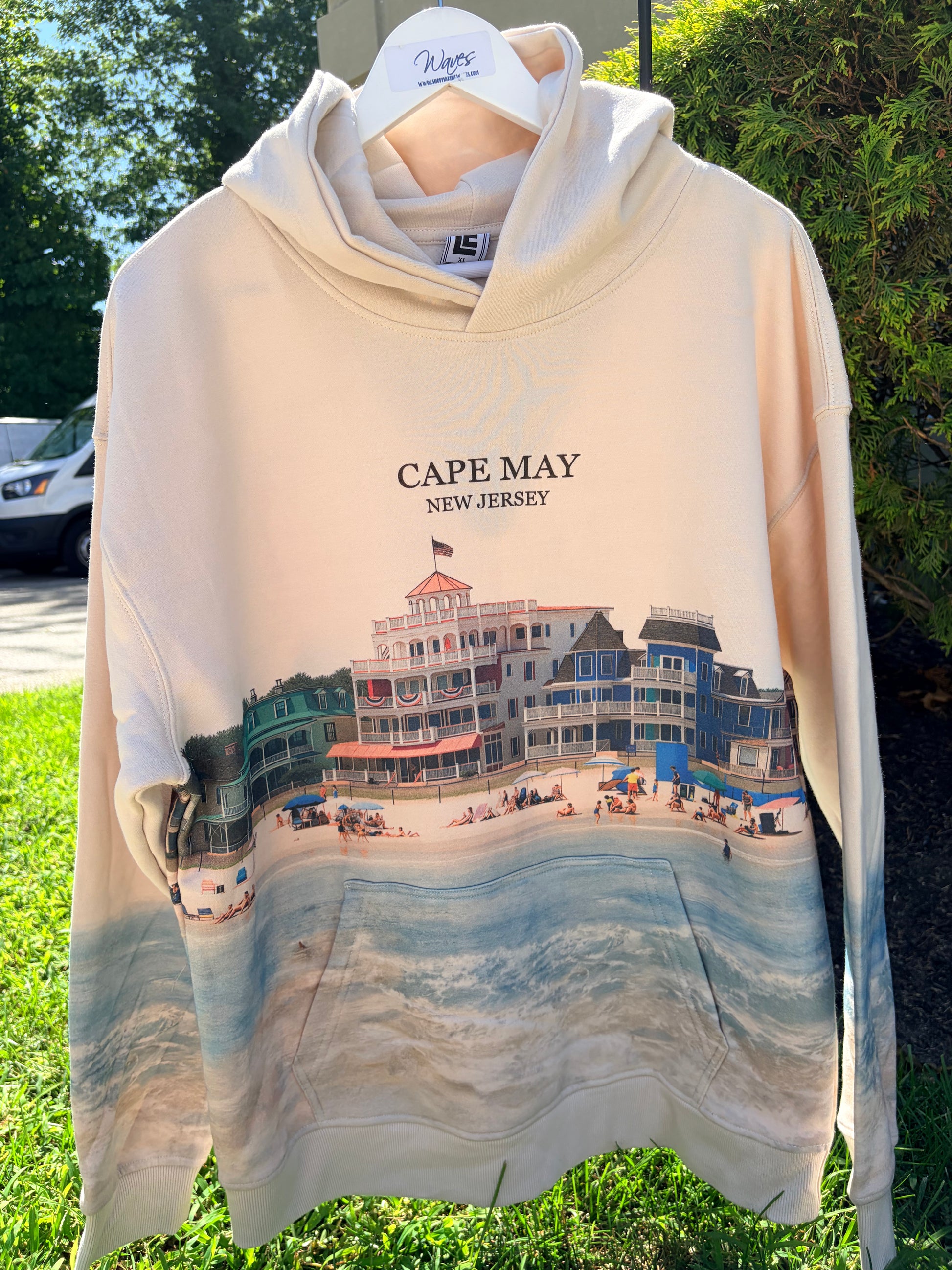 A cream-colored hoodie with a 'Cape May New Jersey' beach boardwalk graphic, hanging on a white hanger outdoors on grass with trees and a vehicle in the background.